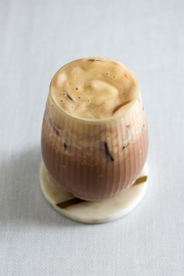 A close-up of an iced caramel chocolate cold brew in a textured glass, sitting on a marble coaster with a smooth caramel foam topping