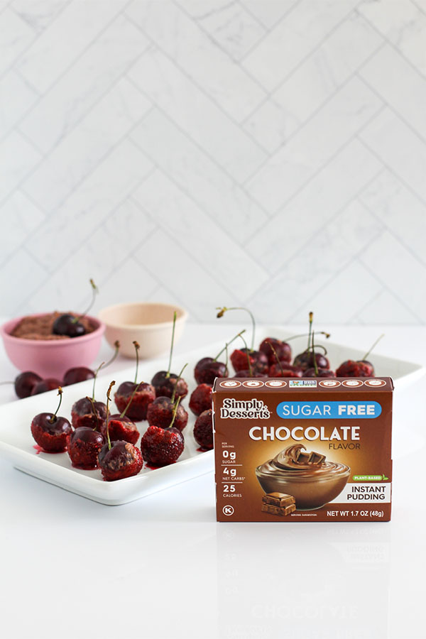 Simply Desserts Sugar Free Chocolate Pudding box displayed alongside a platter of Chocolate Boozy Cherry Bites, a bowl of pudding, and fresh cherries on a white surface