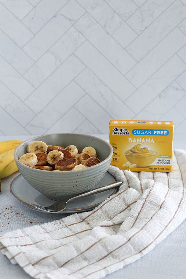 A cozy breakfast setting featuring a bowl of banana griddle bites, fresh bananas, and Simply Desserts Sugar Free Banana Pudding on a soft kitchen towel