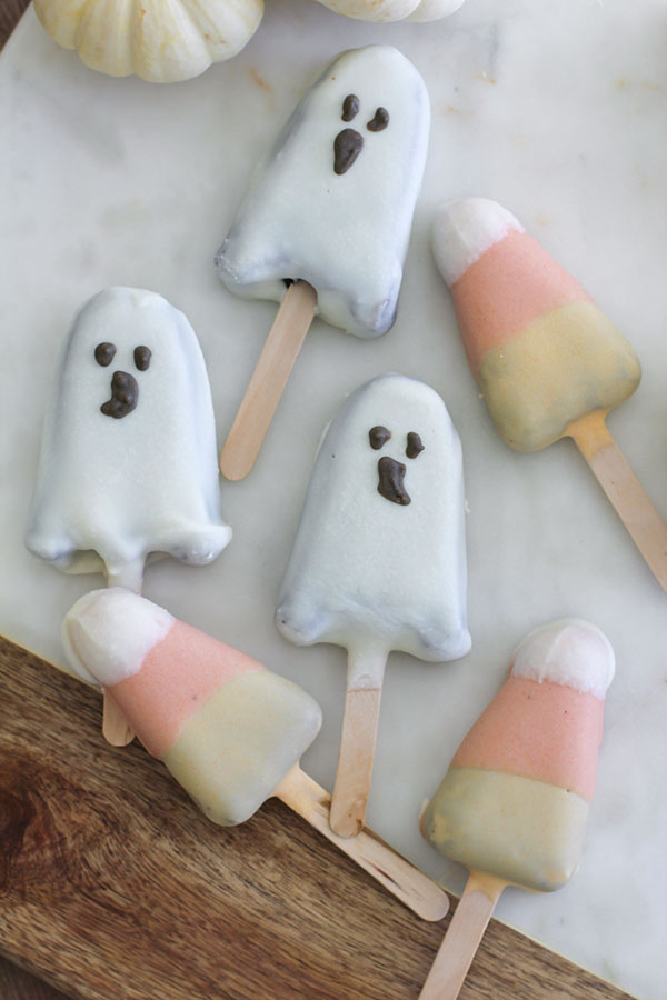 Assorted Halloween cake pops shaped like ghosts and candy corn, decorated with white, orange, and yellow chocolate, displayed on a marble and wood surface with mini white pumpkins in the background