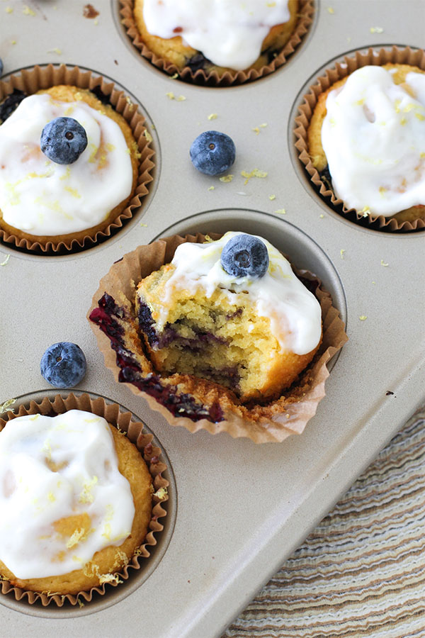 close-up of lemon blueberry muffins in a muffin tin, topped with white frosting and a single blueberry, one muffin bitten to reveal the inside