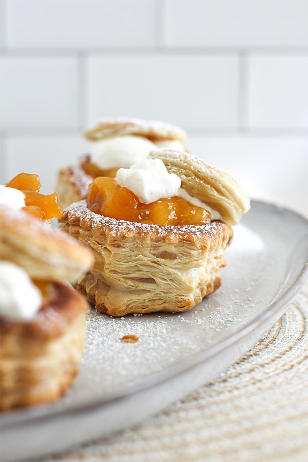 Close-up of peach pie pastry cups on a serving plate, filled with peach chunks, topped with whipped cream and a dusting of powdered sugar
