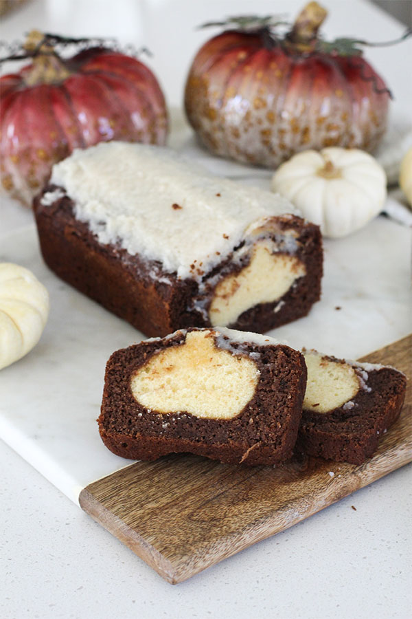 Chocolate loaf cake with white frosting, sliced to reveal a hidden pumpkin shape, surrounded by decorative pumpkins — perfect for Halloween or fall desserts
