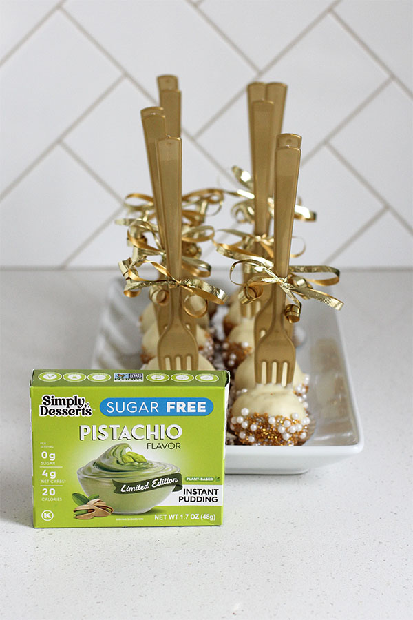 Simply Desserts Sugar Free Pistachio Pudding box displayed in front of a tray of Pistachio White Chocolate Cake Bites with gold forks and ribbons
