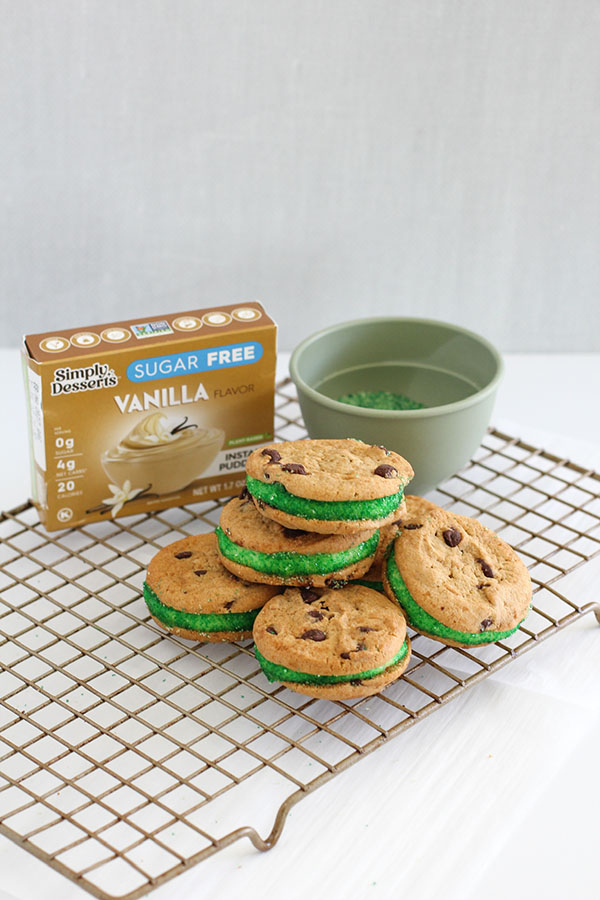 Pudding Cookie Sandwiches - A batch of pudding cookie sandwiches with green sprinkles, displayed on a cooling rack with a Simply Desserts Vanilla Pudding box and a green bowl of sprinkles