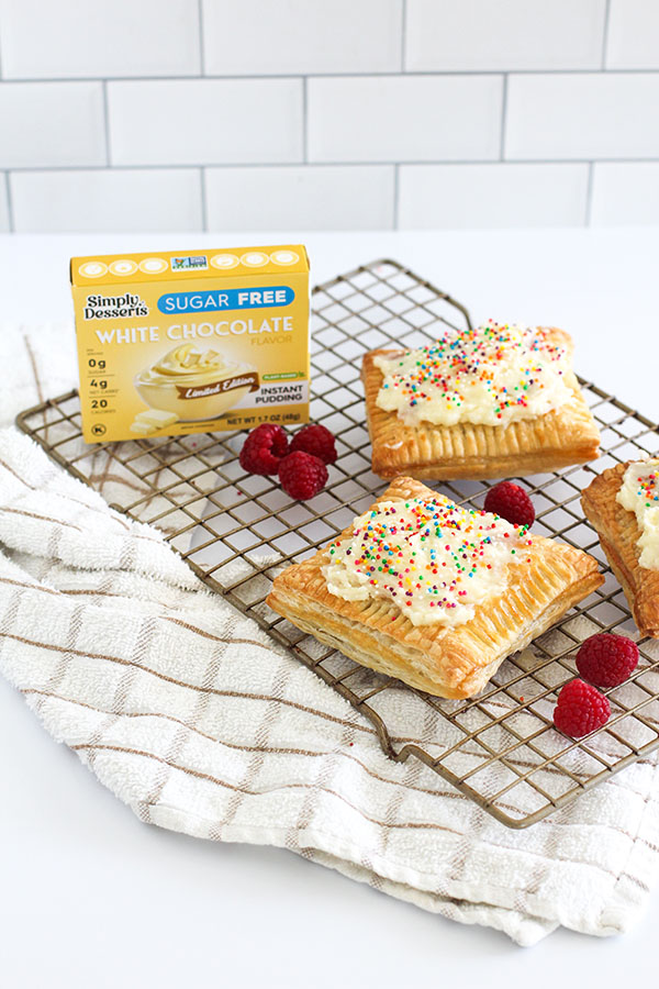Raspberry White Chocolate Pastry Tarts with pudding and sprinkles on a cooling rack, with fresh raspberries and a pudding box