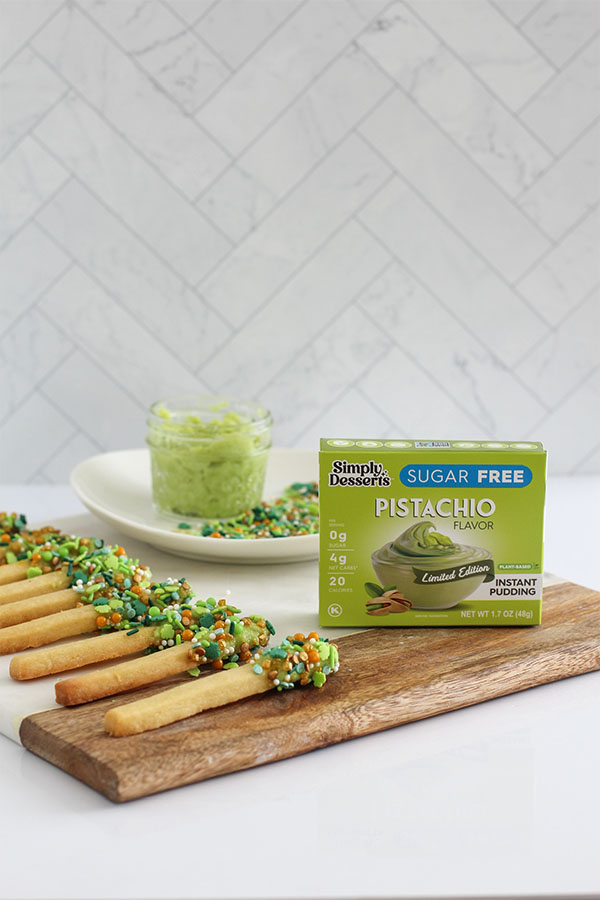 Pistachio shortbread cookie sticks displayed on a wooden board with Simply Desserts Pistachio Pudding box