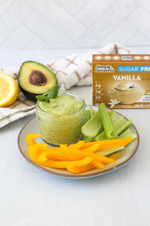 Green Goddess Dip served in a glass jar on a plate with celery and yellow peppers, Simply Desserts vanilla pudding box in background