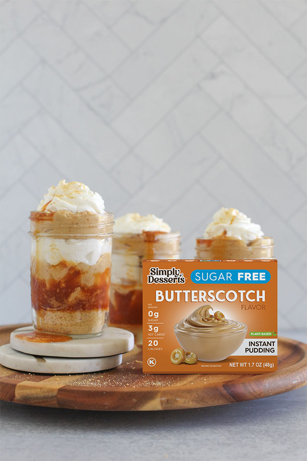 Three salted caramel butterscotch trifles in jars with a Simply Desserts Sugar Free Butterscotch Pudding box in front, set on a wooden tray with a tiled background