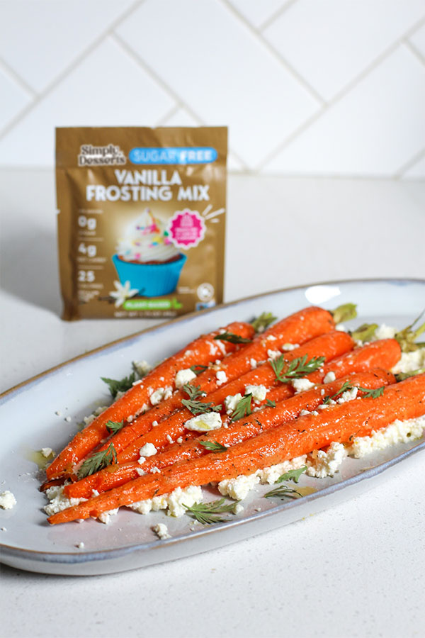 A tray of roasted carrots on ricotta with fresh herbs, positioned in front of a standing Simply Desserts Vanilla Frosting Mix packet on a kitchen counter