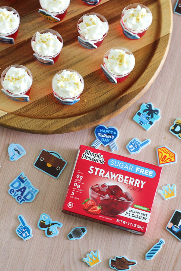 Overhead view of strawberry jel shots topped with cream and golden sprinkles on a wooden platter, surrounded by Father's Day-themed stickers