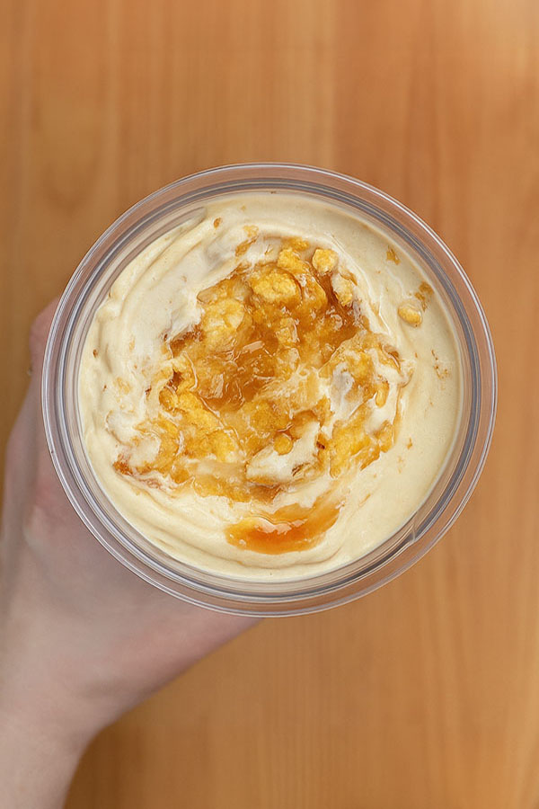 Top-down view of a hand holding a glass bowl of peanut butter caramel crunch protein ice cream, topped with golden caramel sauce and crushed cornflakes