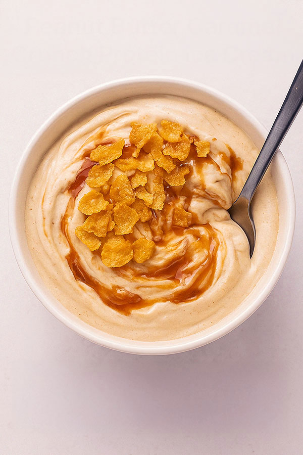 Top-down view of a hand holding a bowl of peanut butter caramel crunch protein ice cream, topped with golden caramel sauce and crushed cornflakes