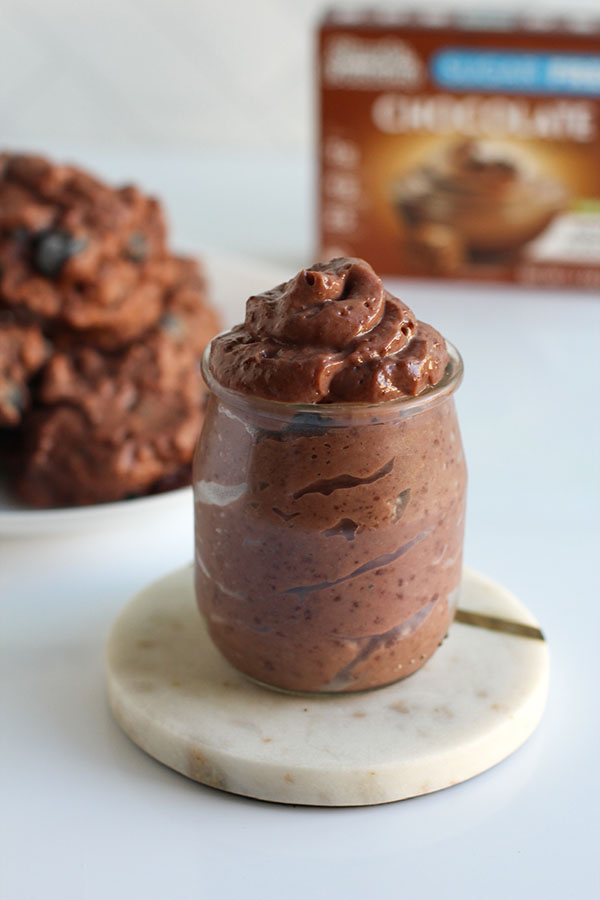 Glass jar filled with creamy Simply Desserts Sugar Free Chocolate Pudding on a marble coaster, with frozen blueberry clusters blurred in background