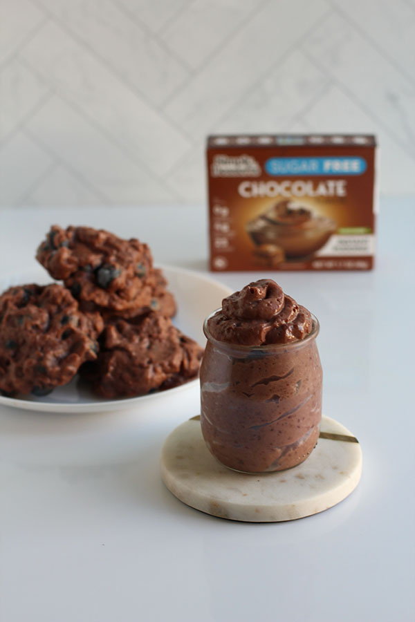 Creamy jar of Simply Desserts Chocolate Pudding in front of a plate of frozen clusters and the product box