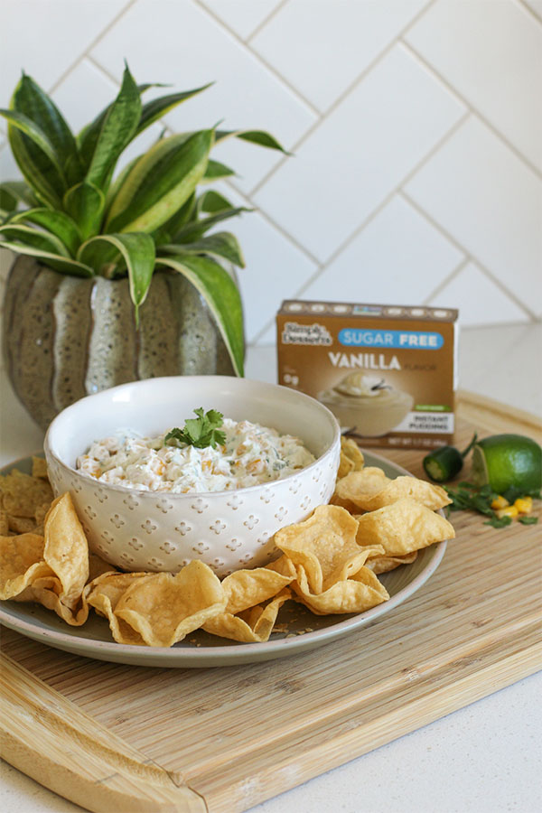 Spicy Pudding Corn Dip served in a white patterned bowl on a plate with tortilla chips, pudding mix box, and a plant in the background
