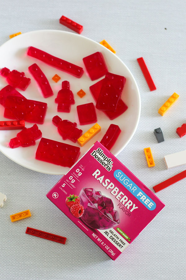 Plate of raspberry-flavored jelly Lego bricks and mini-figure shapes next to a Simply Desserts Sugar Free Raspberry Jel Dessert box, surrounded by colorful Lego pieces