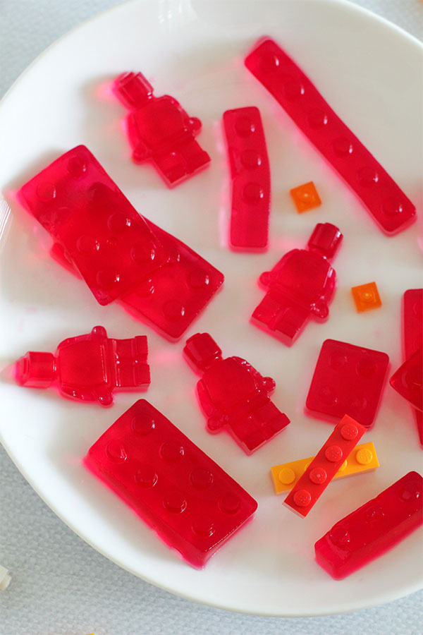 Raspberry-flavored jelly molded into Lego bricks and mini-figure shapes on a white plate, with small orange and yellow Lego pieces for decoration.
