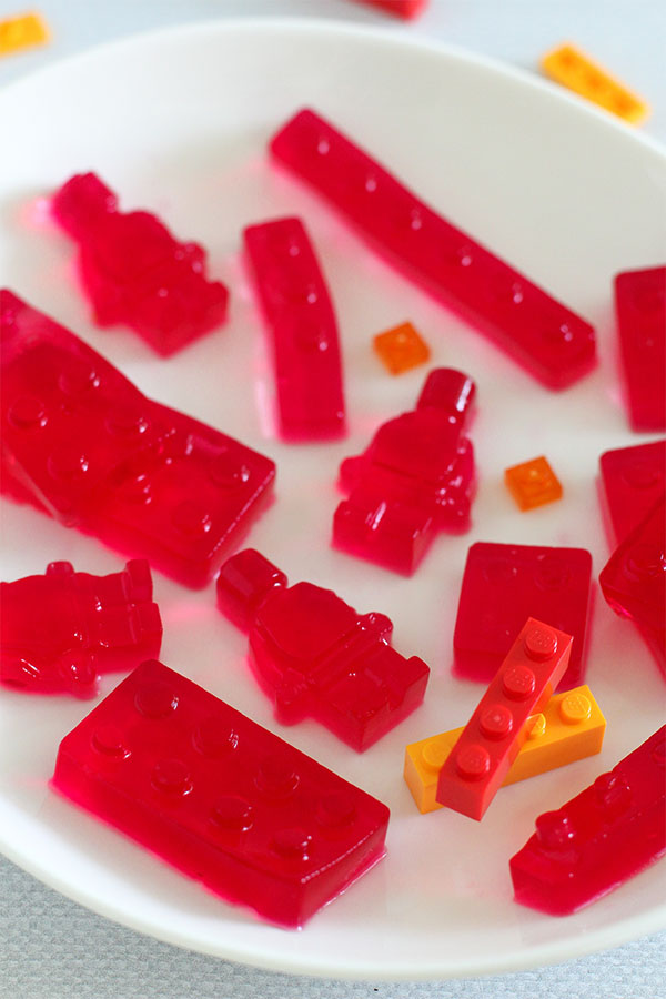 Close-up of raspberry-flavored jelly molded into Lego bricks and mini-figure shapes, served on a white plate with small plastic Lego pieces for decoration