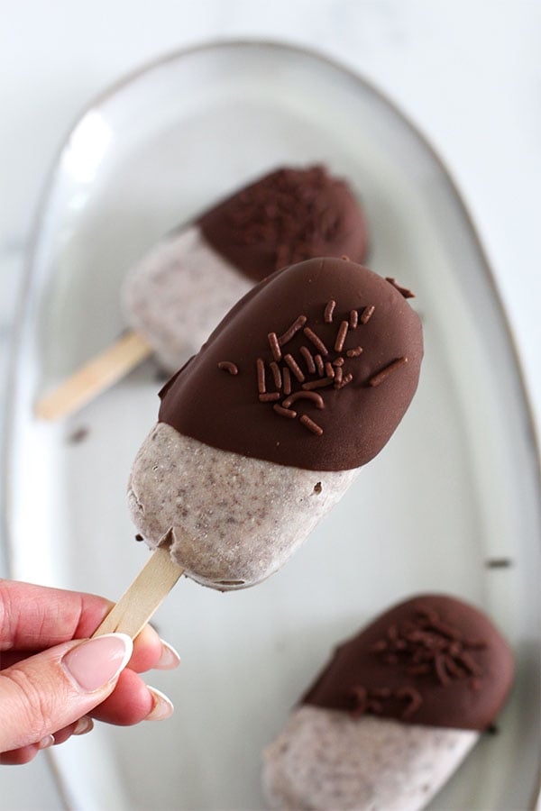 Hand holding a cookies and cream pudding pop dipped in dark chocolate and topped with chocolate sprinkles, with more pops on a plate in the background