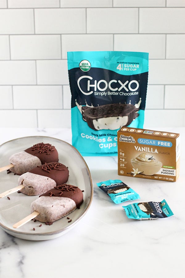 Cookies and cream pudding pops dipped in dark chocolate with sprinkles, served on a plate next to Chocxo Cookies & Cream Cups packaging and Simply Desserts Sugar Free Vanilla Pudding Mix box on a white countertop