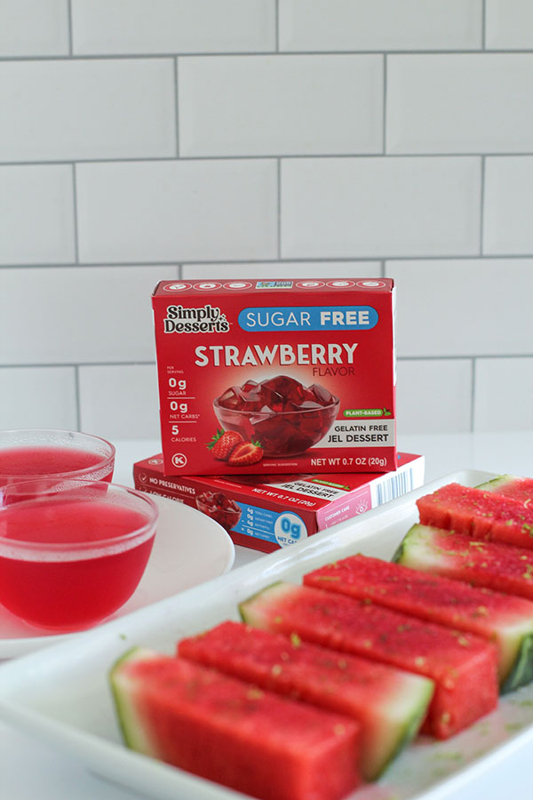 Simply Desserts Sugar Free Strawberry Jel cups and fresh watermelon spears with lime zest displayed on a counter, with product boxes in the background against a white tiled wall