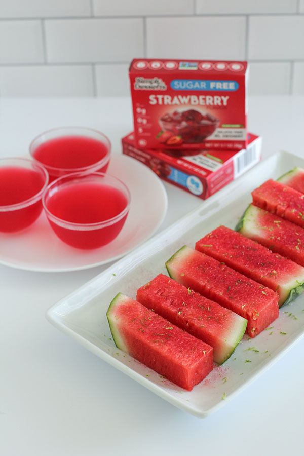 Plate of vibrant watermelon spears topped with lime zest next to three cups of Simply Desserts Sugar Free Strawberry Jel, with product boxes in the background.