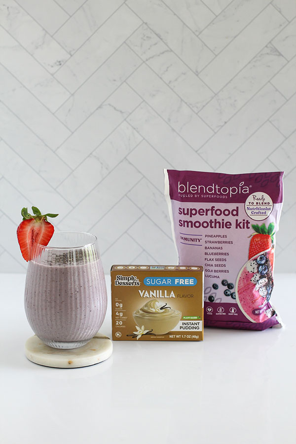 Glass of purple immunity smoothie garnished with a fresh strawberry, displayed alongside a box of Simply Desserts sugar-free vanilla instant pudding and a Blendtopia immunity superfood smoothie kit package