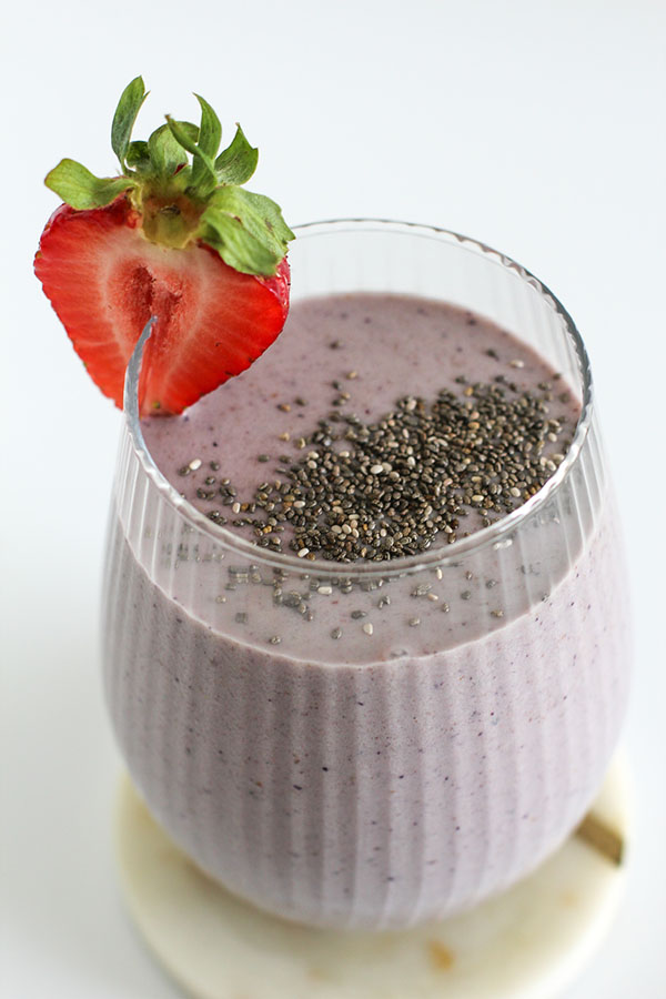 Close-up view of a purple smoothie topped with chia seeds and garnished with a halved fresh strawberry, served in a ribbed glass on a marble coaster.
