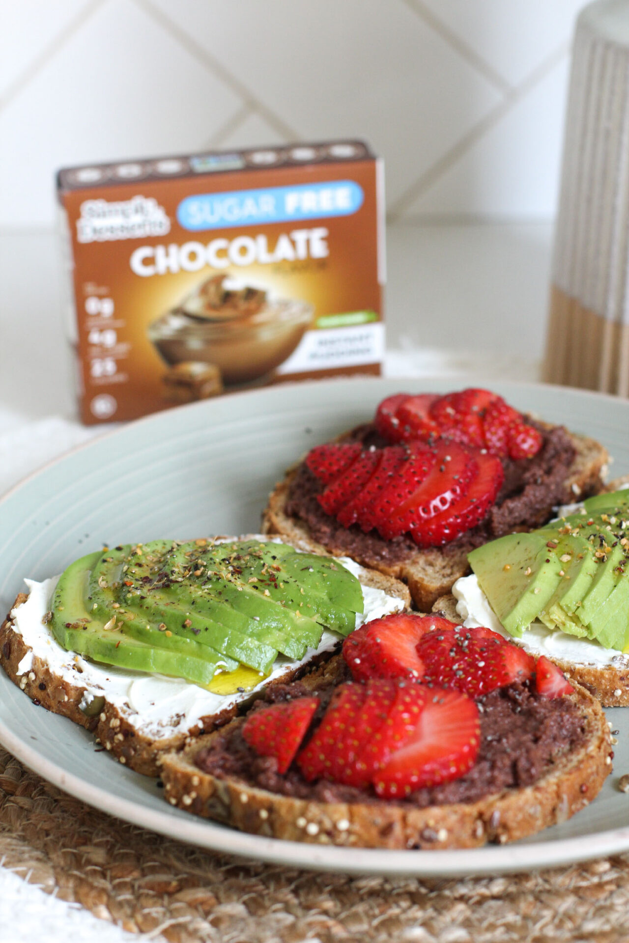 Plate of sweet and savory breakfast toasts topped with avocado, cream cheese, strawberries, chia seeds, and Simply Desserts Sugar Free Chocolate Pudding, with product box and vase in the background