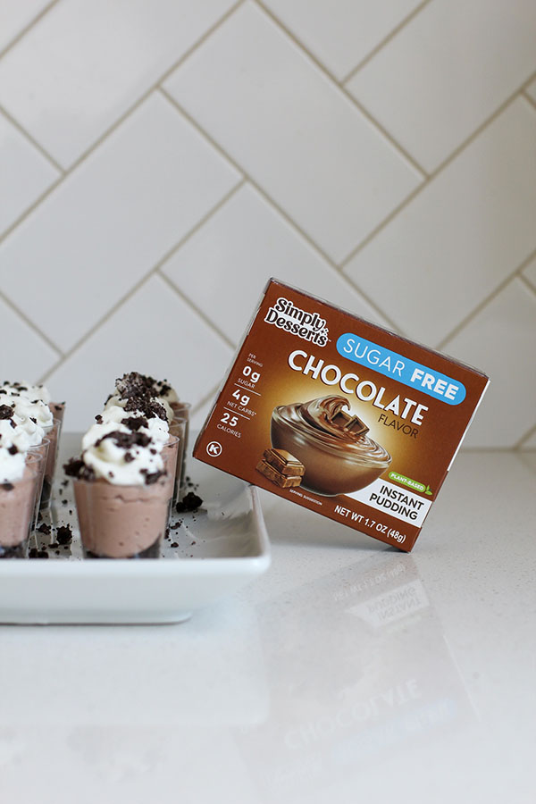 Simply Desserts Sugar Free Chocolate Pudding box displayed beside mini chocolate cheesecake parfaits in small cups, layered with cookie crumbs, chocolate filling, and whipped cream topping