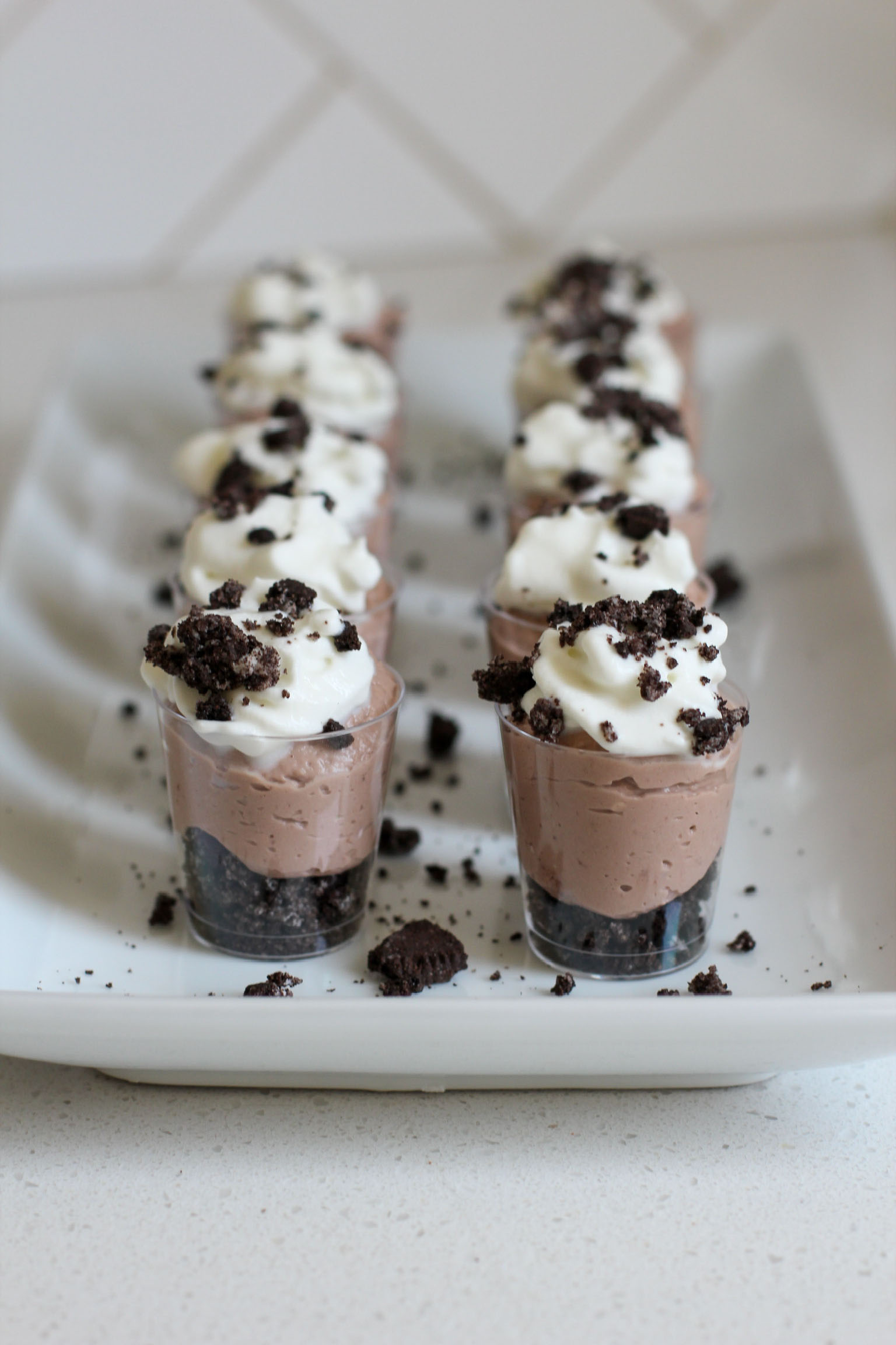 Row of mini chocolate cheesecake parfaits in clear cups with a cookie crumb base, creamy chocolate filling, whipped cream topping, and sprinkled cookie pieces on a white tray