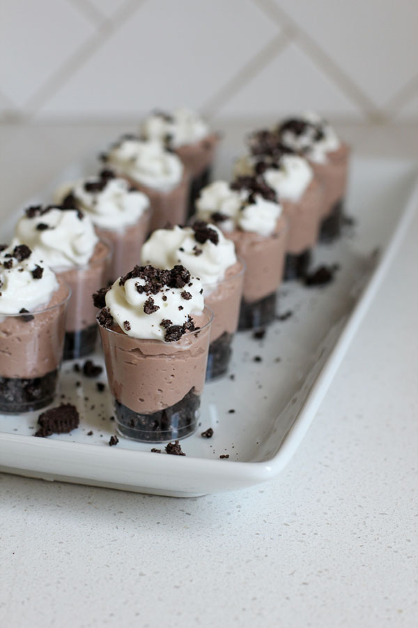 Tray of mini chocolate cheesecake parfaits with a crushed cookie base, smooth chocolate filling, whipped cream topping, and sprinkled cookie crumbs, arranged in neat rows