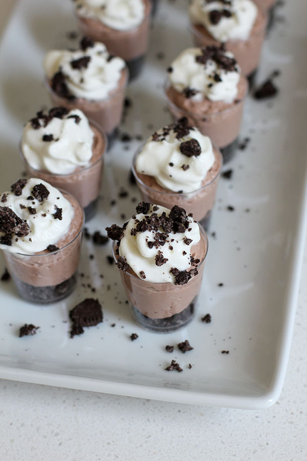 Mini chocolate cheesecake parfaits served in small clear cups, topped with whipped cream and sprinkled with crushed chocolate cookies, arranged neatly on a white tray