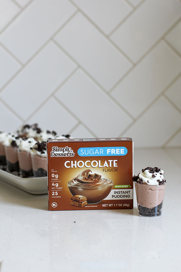 Simply Desserts Sugar Free Chocolate Pudding box displayed on a counter with a mini chocolate cheesecake parfait in front and a tray of additional parfaits in the background