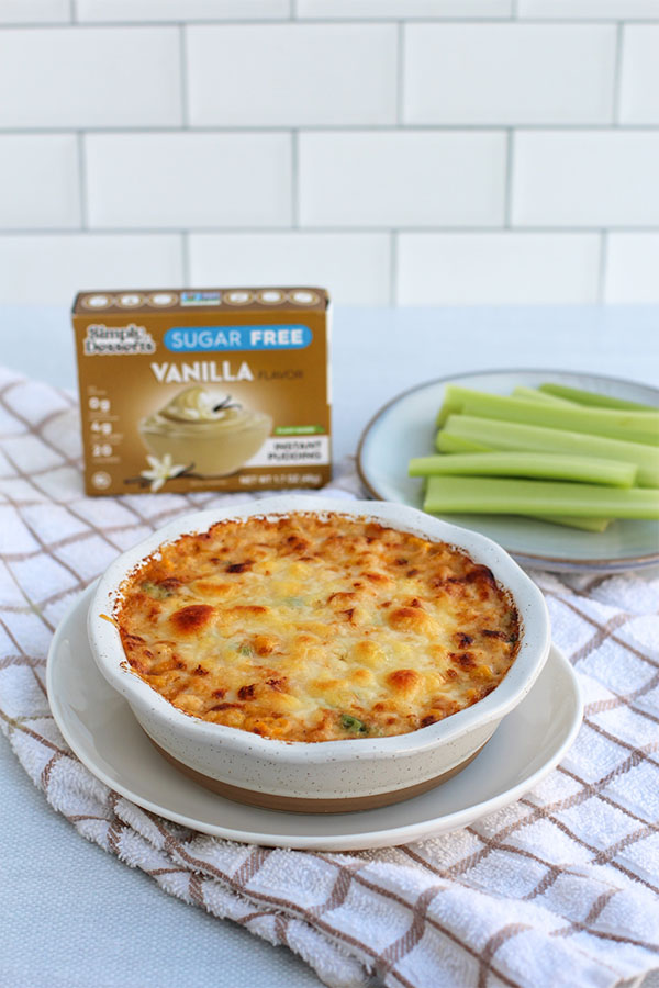 Spicy Corn Pudding Dip baked with cheddar cheese in a white dish, served with celery sticks and Simply Desserts Sugar Free Vanilla Pudding box in the background