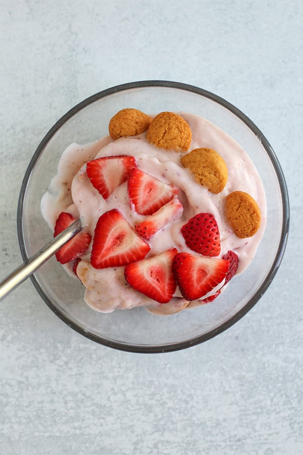 Top view of strawberry shortcake ice cream in a glass bowl, topped with fresh strawberry slices and whole vanilla wafers, with a spoon inside.