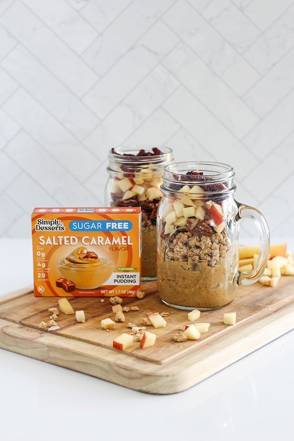 Two glass jars layered with Simply Desserts Salted Caramel Pudding, granola, diced apples, and dried cranberries on a wooden board beside a Salted Caramel pudding box.