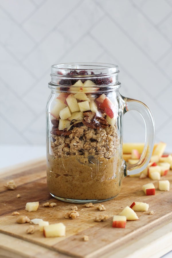 Close-up of a single jar showing layers of salted caramel pudding, crunchy granola, diced apple, and cranberries on a wooden board