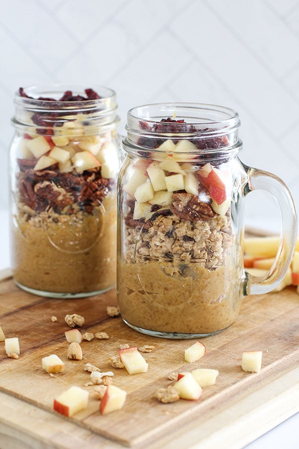 Two caramel pudding parfaits in glass mugs topped with apples and cranberries, surrounded by apple chunks and granola on a board