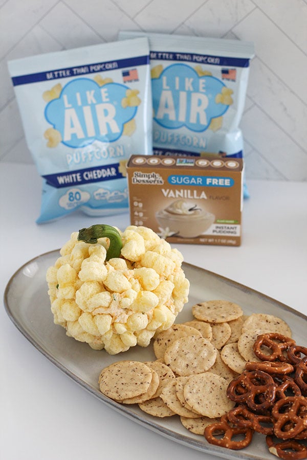 Cheeseball appetizer plated with pretzels and crackers, showing Simply Desserts Vanilla Pudding and Like Air Puffcorn packaging in the background for a cozy fall party setup