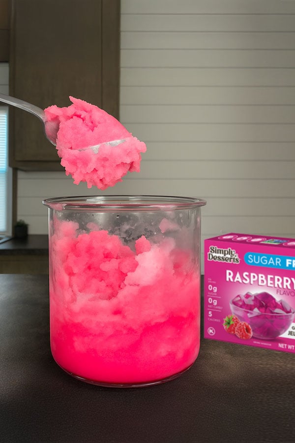 Ninja Creami Pint with pink raspberry lemonade sorbet, with a spoonful lifted above the cup and a Simply Desserts Raspberry Jel mix box in the background on a kitchen counter