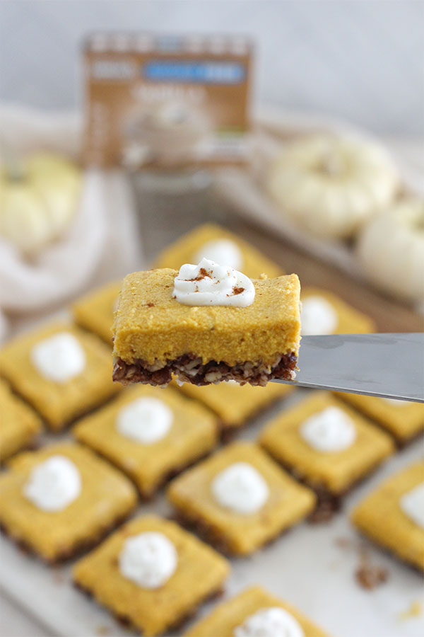 Close-up of a creamy pumpkin pie bar topped with whipped cream, held above a tray of no-bake pumpkin squares