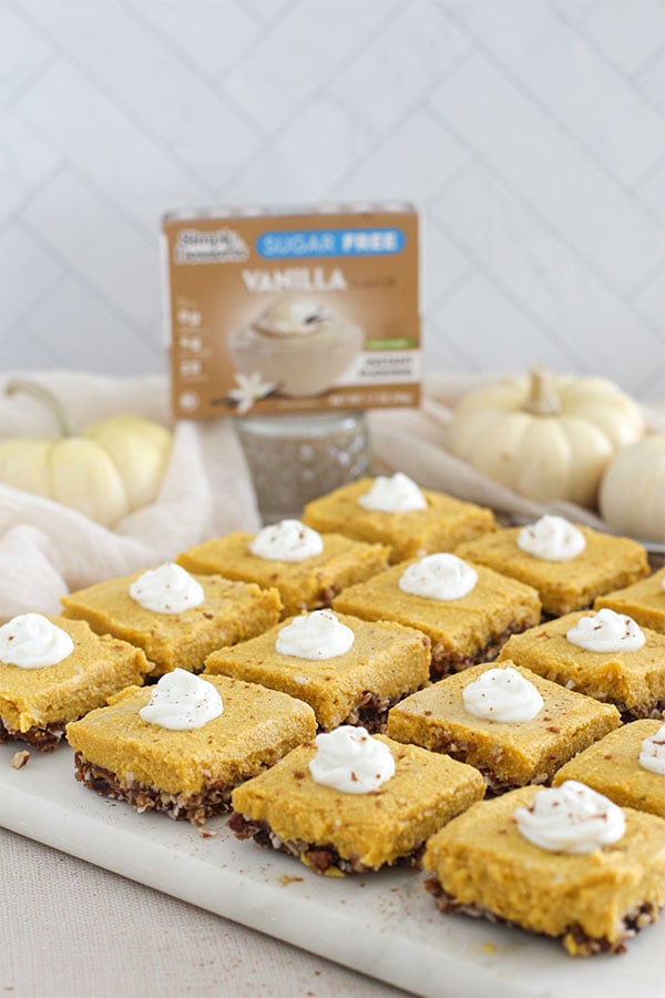 Rows of pumpkin pie bars topped with whipped cream, with Simply Desserts Vanilla Pudding box blurred in the background
