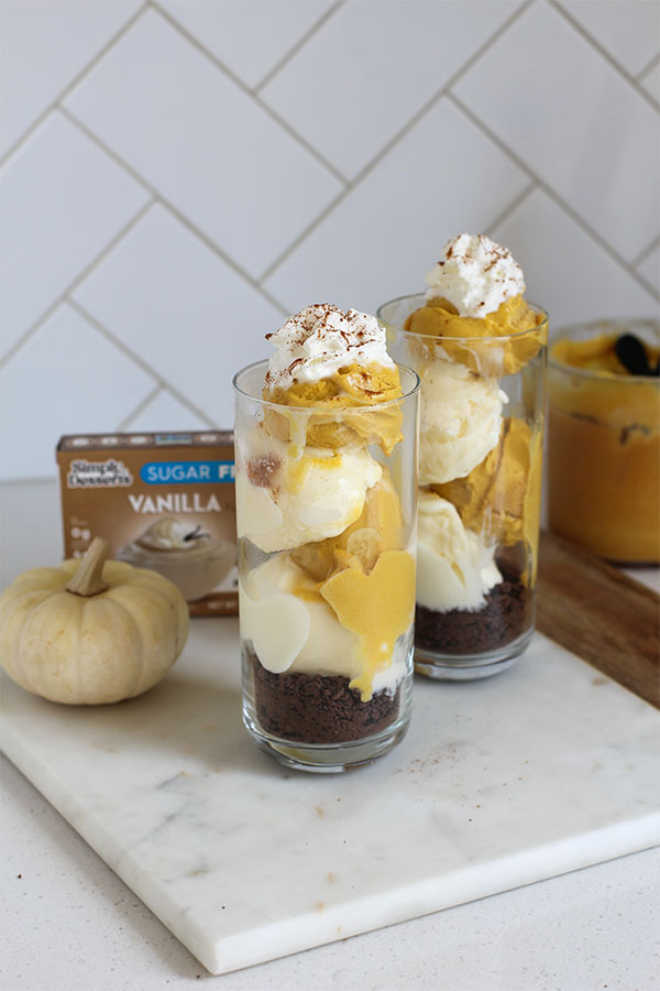 Two tall glasses filled with layers of pumpkin and vanilla pudding topped with whipped cream and cinnamon, set on a marble surface beside a Simply Desserts Sugar-Free Vanilla Pudding box and a small white pumpkin