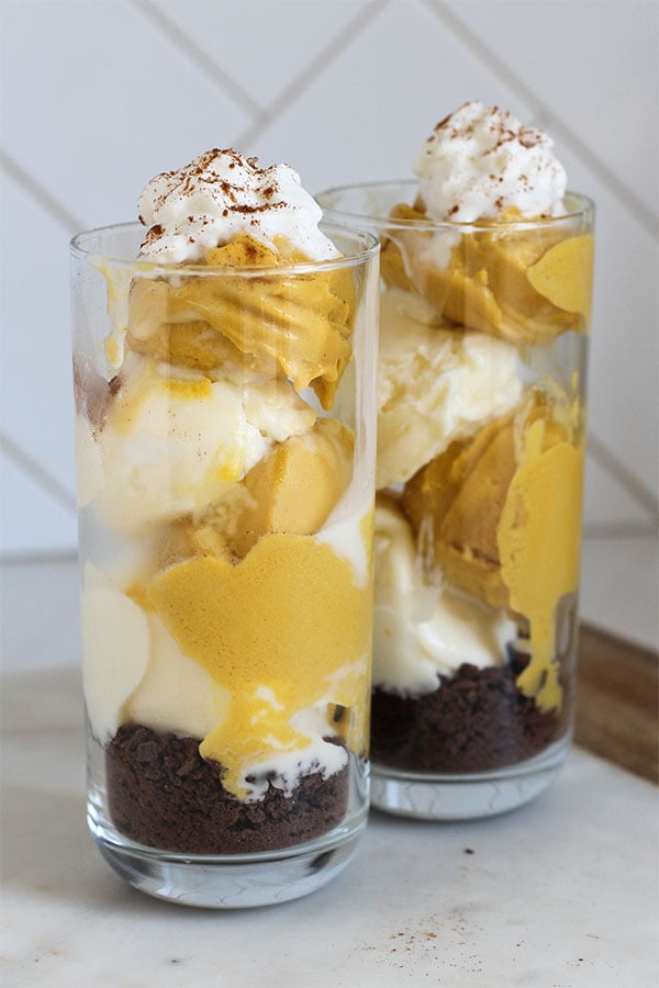 Close-up of two dessert glasses filled with layers of pumpkin and vanilla pudding over a chocolate crumb base, topped with whipped cream and sprinkled with cinnamon for a cozy fall treat