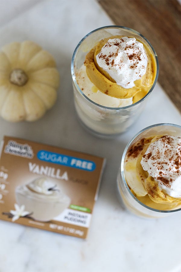 Overhead shot of two glasses filled with layers of pumpkin and vanilla pudding topped with whipped cream and cinnamon, next to a Simply Desserts Sugar-Free Vanilla Pudding box and a small white pumpkin on a marble counter