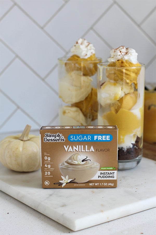 Simply Desserts Sugar-Free Vanilla Pudding box displayed on a marble board with two tall pumpkin and vanilla pudding sundaes topped with whipped cream and cinnamon in the background, styled with a small white pumpkin for a cozy fall look
