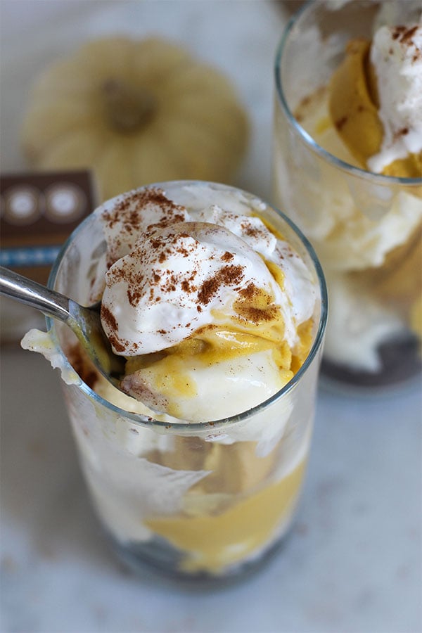 Close-up of a spoon dipping into creamy layers of pumpkin and vanilla pudding topped with whipped cream and sprinkled with cinnamon, showcasing the rich texture of the fall-inspired dessert