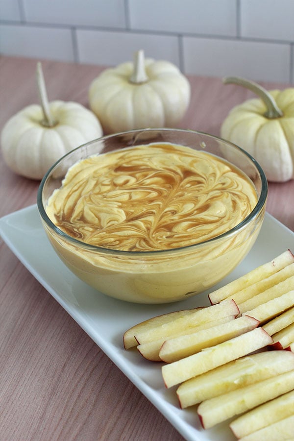 Bowl of pumpkin spice cheesecake dip with caramel swirl served alongside crisp apple slices and white mini pumpkins